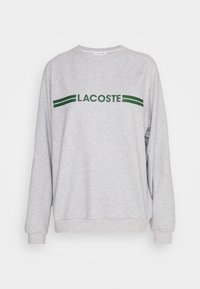 Grynas džemperis su ilgomis rankovėmis, raukšlėtais rankogaliais ir apatine dalimi. Su žaliu "LACOSTE" logotipu ir dviem žaliomis juostelėmis virš jo. Minkšta tekstūra.
