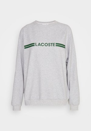 Grijze sweatshirt met lange mouwen, ribbelboorden en een ribbelhem. Heeft een groen "LACOSTE" logo met daarboven twee groene strepen. Zachte textuur.
