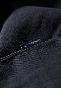 Tessuto in denim scuro con una texture liscia, caratterizzato da un'etichetta blu cucita con la scritta "LINDBERGH". Primo piano sul dettaglio dell'etichetta.