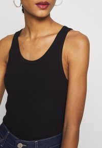 Donna che indossa un top senza maniche rigato nero e jeans blu scuro, con un grande orecchino a cerchio argento visibile sull'orecchio sinistro.