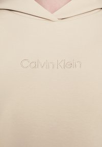 Beige mikina s kapucí, jemná na dotek, s vyšitým logem "Calvin Klein" na přední straně. Má stažitelnou kapuci a minimalistický design.