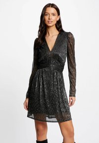 Zwarte jurk met lange doorschijnende mouwen, diepe V-hals, textiele stof met metalen draden en een gegatherde taille.