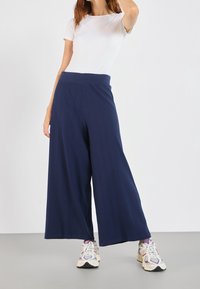 Pantalon large bleu marine avec tissu texturé et taille haute, porté avec une chemise blanche et des baskets colorées.