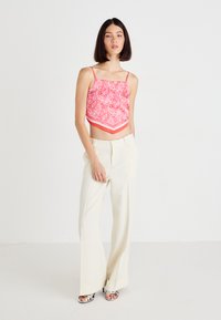Ružičasti paisley crop top s tankim trakama, u kombinaciji s kremastim širokim hlačama i srebrnim sandalama s trakama, postavljen na jednobojnoj pozadini.