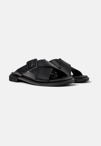 Black leather sandals featuring cross-strap design, rectangular buckle accent, and flat rubber sole for stability and comfort.