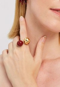 Dwa przerośnięte pierścienie o błyszczących wykończeniach: jeden czerwony, a drugi złoty. Pokazana jest neutralna ręka, która delikatnie dotyka policzka.