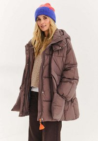 Brązowa pikowana kurtka puchowa z kapturem i pomarańczowym suwakiem; zestawiona z beżowym swetrem i fioletowo-pomarańczową dzianą czapką.