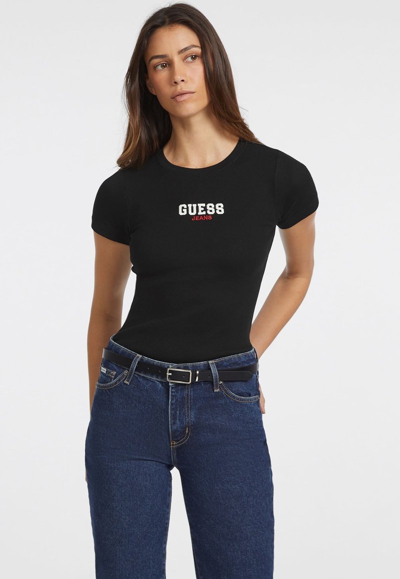 T-shirt noir ajusté à manches courtes, arborant un logo "GUESS JEANS" en blanc et rouge sur la poitrine, associé à un jean en denim bleu et une ceinture noire.