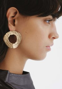 Pendientes circulares en tono dorado con un diseño texturizado y tejido, y un centro abierto. Gran forma audaz sobre un modelo contra un fondo blanco.