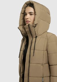 Tawny pufferjack met hoge kraag en oversized capuchon, voorzien van zwarte trekkoorden en een gevoerde textuur, ontworpen voor warmte en comfort.