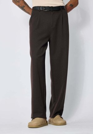 Homme portant un pantalon marron foncé à jambes larges, ceinture noire, chaussures beige sans lacets, debout devant un fond clair uni avec les mains dans le dos.