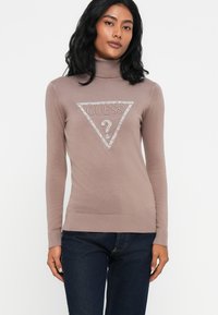 Donna che indossa un maglione a collo alto mauve con logo Guess impreziosito e jeans blu scuro, in piedi davanti a uno sfondo bianco semplice.