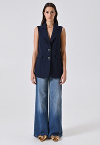 Marineblaues ärmelloses Blazer mit zwei Fronttaschen und silbernen Beschlägen, kombiniert mit weiten hellblauen Jeans und Schuhen mit goldenen Schleifen-Details.