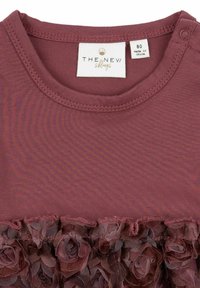 Bordo majica s mekanom pamučnom teksturom, s ukrasnim cvjetnim čipkastim volanom. Okrugli izrez i detalj s kopčanjem na ramenu. Vidljiva oznaka veličine.