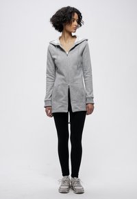 Svijetlosiva majica s patentnim zatvaračem i bijelim rubom na kapuljači, dva prednja džepa i dugim krojem, uparena s crnim tajicama i sivim tenisicama.