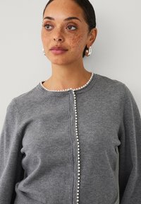 Grå strikket cardigan med perle-accenter langs den runde halsudskæring og frontåbning; blød tekstur og tætsiddende design.