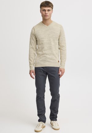 Pull en tricot beige avec un col en V et des poignets côtelés, associé à un pantalon slim gris foncé et des baskets beiges avec des accents clairs.