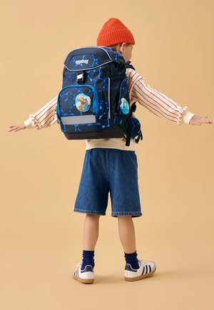 Child wearing a large blue and black patterned backpack with dinosaur patches, orange beanie, striped sweater, denim shorts, and sneakers, arms outstretched.