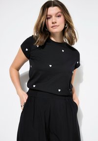 Černé tričko s rozptýlenými bílými srdíčky, krátké rukávy a kulatý výstřih. V kombinaci s volnými černými kalhotami.