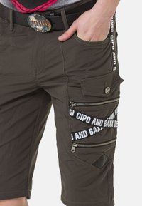 Cargo-Shorts in dunkeloliv mit mehreren Taschen, Reißverschlüssen und weißen Markierungsdetails. Hergestellt aus leichtem Stoff mit einer glatten Textur.