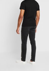 Hombre con camiseta negra, jeans negros desteñidos y zapatillas blancas de pie contra un fondo liso claro, de espaldas.