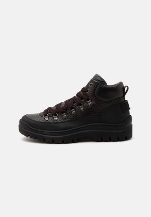 Bota de senderismo negra de piel con caña alta, suela gruesa de goma, ojales metálicos y cordones marrón oscuro mostrada de lado sobre un fondo blanco.