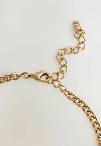 Goldene Metallkette mit ovalen Gliedern, ausgestattet mit einem Karabinerverschluss und einem rechteckigen Beschlag mit einem kleinen Steinelement.
