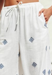 Vita linnebyxor med dragsko i midjan, sidofickor och blå broderade diamantmönster på tyget. Lätta och strukturerade.