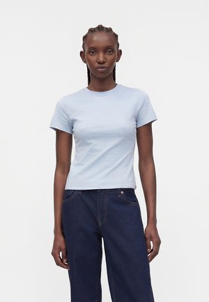 Γυναίκα με πλεξουδάκια, που φορά ένα ανοιχτό μπλε κοντομάνικο μπλουζάκι και σκούρο τζιν ψηλόμεσο, στέκεται μπροστά σε ένα απλό λευκό φόντο.