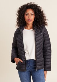 Giacca nera imbottita con zip frontale, cappuccio e cuciture diagonali. Indossata sopra una maglietta chiara e jeans blu, con dettagli di tasche discreti.