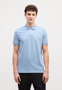 Gaiši zilā polo krekls ar īsām piedurknēm, mīksta tekstūra un neliels izšūts logotips uz krūtīm. Tam ir standarta apkakle un pogu rindas.