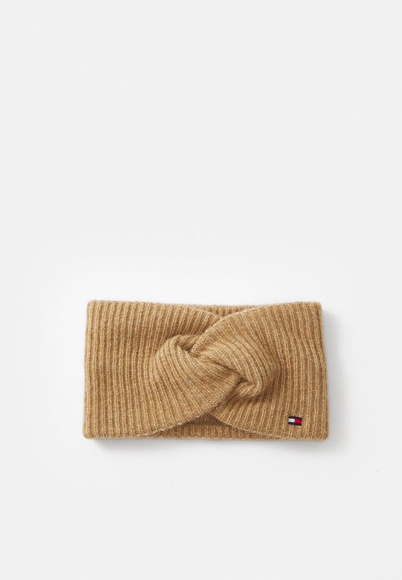 Knit headband in light brown with a twisted design at the front and a small logo tag featuring red, white, and blue accents.