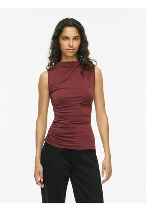 Femme aux cheveux bouclés portant un haut bordeaux froncé sans manches et un pantalon noir, debout devant un fond blanc uni.