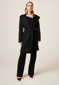 Cappotto di lana nero con ampio colletto, vita cintata e due tasche frontali. Abbinato a pantaloni neri a gamba larga e décolleté rossi a punta.