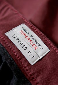 Bordeaux stoflabel met witte en rode tekst die "VRIJHEID VAN BEWEGING SUPERFLEX TAPERSNIT" leest, omringd door een zwarte stikrand.