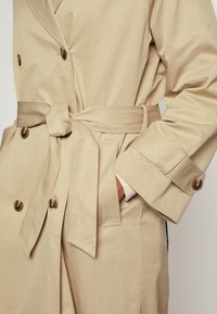 Beige trenchcoat met een tailleceintuur, dubbele knopenrij en zijzakken. Kenmerkt zich door een gladde textuur en opgevouwen manchetten.