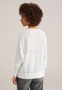 Pull en maille blanche avec rayures horizontales, coupe décontractée, poignets et ourlet côtelés, tissu texturé et manches larges. Porté avec un jean gris.