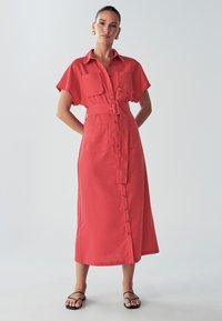 Czerwona sukienka do kolan z krótkimi rękawami, z pasem w talii, dwiema kieszeniami na piersi i guzikami na przodzie. Gładka tekstura materiału.