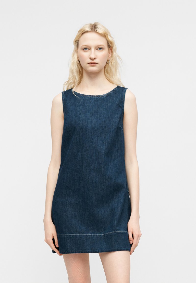 Νεαρή γυναίκα με ελαφρώς κυματιστά ανοιχτά μαλλιά, φορώντας ένα αμάνικο σκούρο μίνι φόρεμα από τζιν, στέκεται μπροστά σε ουδέτερο φόντο.