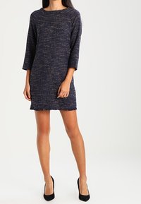 Modelka w granatowej, textured sukience z trzema ćwierć rękawami, stojąca na białym tle, w połączeniu z czarnymi, wysokimi obcasami.