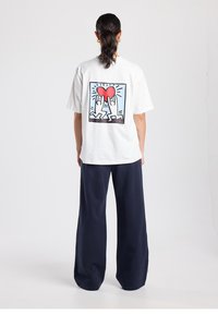 Wit katoenen T-shirt met een grote afbeelding op de achterkant van twee handen die een rood hart vasthouden en zonnestralen, gecombineerd met navy wijde broekspijpen.