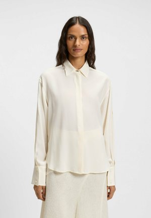 Donna con lunghi capelli scuri che indossa una camicia chiara a maniche lunghe e bottoni, abbinata a una gonna leggera e texture, di fronte a uno sfondo semplice.