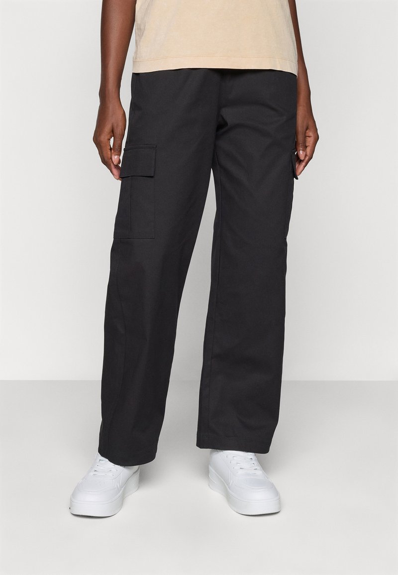 Pantaloni cargo neri con una vestibilità rilassata, dotati di tasche laterali e tessuto leggero. Abbinati a sneaker bianche su uno sfondo neutro.