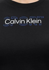 Μαύρο t-shirt με την επιγραφή "Calvin Klein" σε λευκό κείμενο επάνω σε μεγάλα, περιγραμμικά μπλε γράμματα που σχηματίζουν τη λέξη "SPORT" στην περιοχή του στήθους.