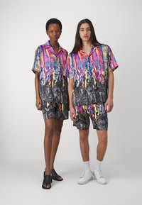 Le camicie a maniche corte e i pantaloni corti colorati presentano un motivo vorticoso in nero, rosa, blu e giallo, realizzati in tessuto leggero.