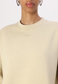Felpa beige con collo tondo e maniche lunghe. Presenta un logo in rilievo discreto sul petto. Realizzata in materiale morbido e testurizzato.