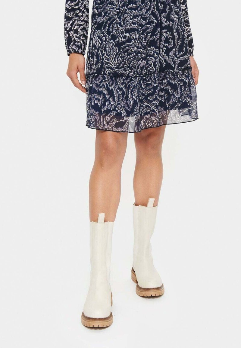 Femme portant une robe à motif floral bleu marine et des bottes mi-mollet blanc cassé avec des semelles beige, se tenant sur un fond blanc sans coutures.