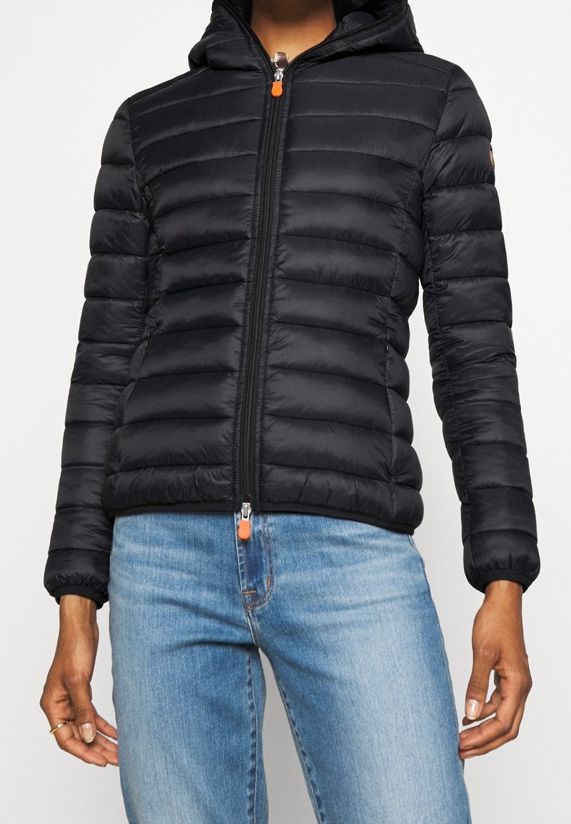 Veste matelassée noire avec capuche et tirettes de fermeture éclair orange portée avec un jean en denim bleu clair sur une personne debout devant un fond blanc.