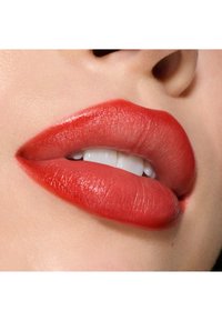 Rossetto rosso brillante applicato sulle labbra, finitura lucida, texture liscia, con bordi definiti. Denti visibili tra le labbra leggermente socchiuse.