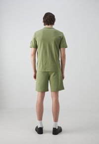 Camisa de manga corta verde oliva y pantalones cortos a juego, hechos de una tela suave, con un corte relajado y costuras mínimas, llevados con zapatos negros.